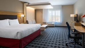 A hotel room with a large bed, a desk with a chair, a small table, a chair, a lamp, and a window with curtains. - TownePlace Suites Columbia West/Lexington (West Columbia)