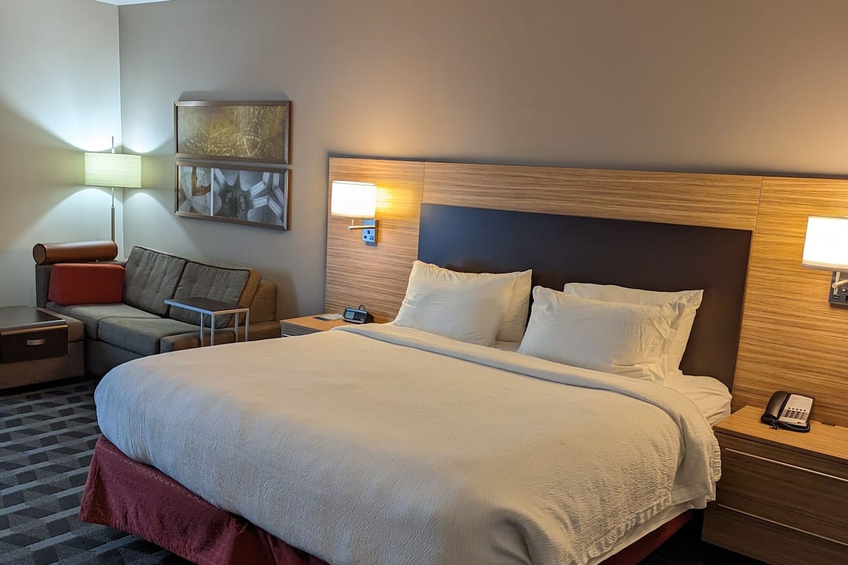 Una habitación de hotel con una cama grande, una mesita de noche, una lámpara, una silla y un teléfono instalado en la pared.