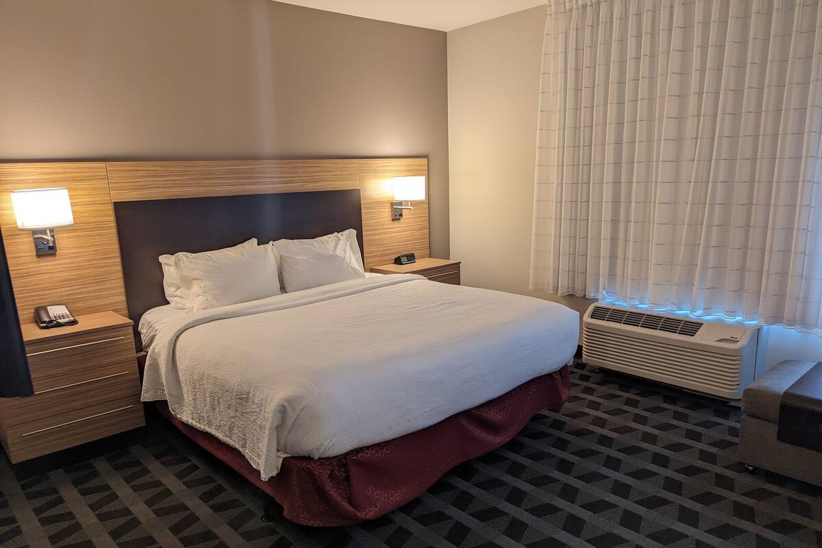 Una habitación de hotel con una cama grande, dos mesitas de noche con lámparas, un calentador y una ventana con cortinas.
