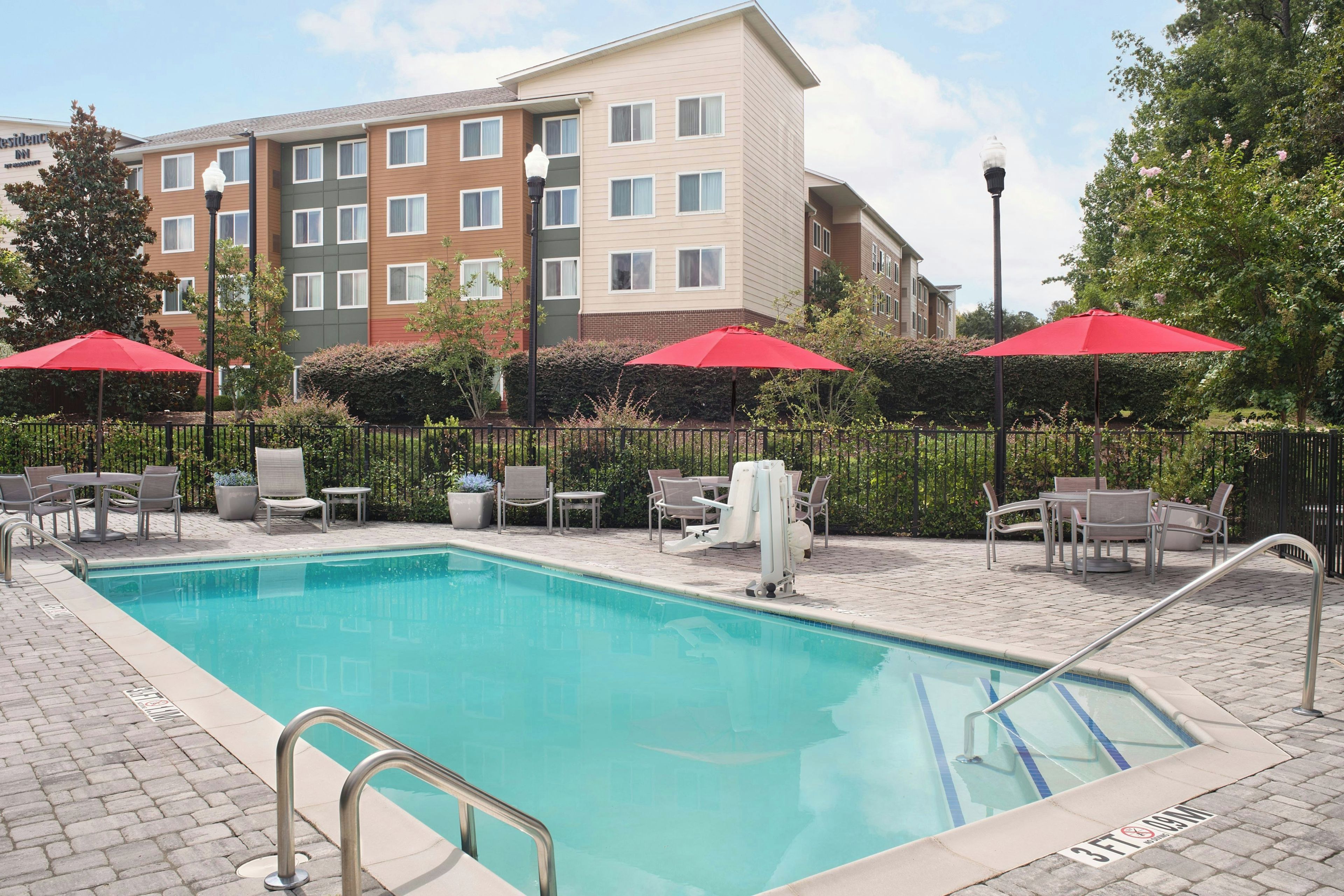A swimming pool with a metal ladder, surrounded by lounge chairs and red umbrellas, with a multi-story building in the background.