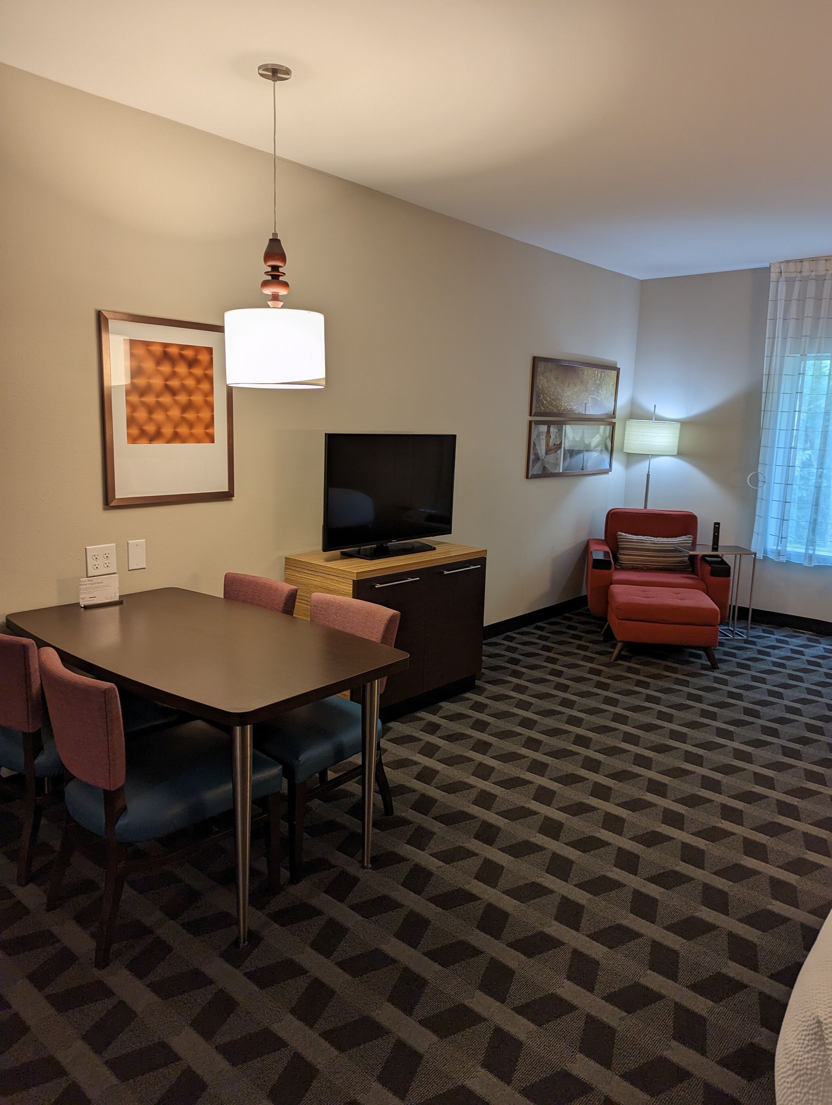 Een hotelkamer met een eettafel, stoelen, een televisie, een rode fauteuil en een kamerbreed tapijt met een patroon.