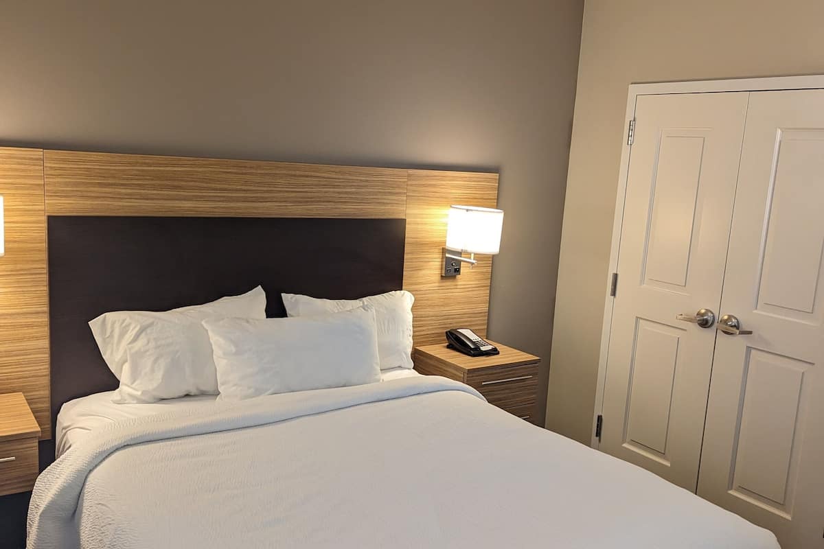 Una cama bien tendida con sábanas blancas, cabecera de madera y una lámpara de noche.