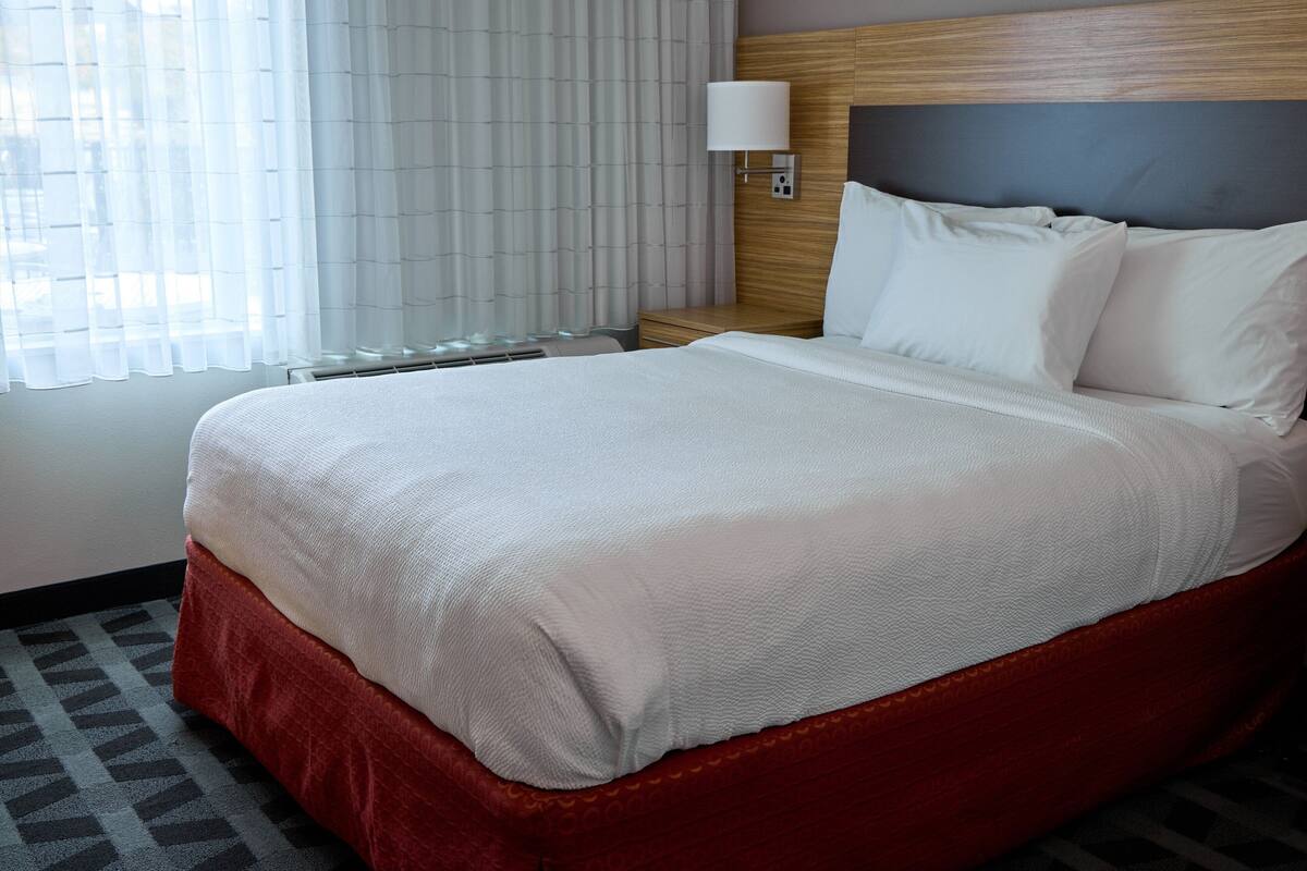 Una habitación de hotel con una cama grande, una mesita de noche con una lámpara y una ventana con cortinas de gasa.