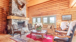 L’intérieur d’une cabane en rondins, avec une cheminée en pierre, des fauteuils en cuir, un tapis rouge et une vue sur les montagnes à travers de grandes fenêtres.