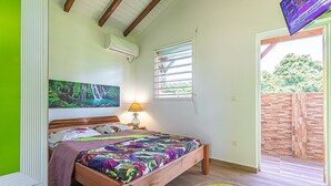 A bedroom with a wooden bed, a painting of a waterfall, a window with blinds, and a view of a brick wall and greenery outside. - Cottage Ramée (Piscine, BBQ, vue sur la mer, montagne, rivière, forêt) (Basse-Terre)