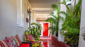 Un pasillo estrecho con piso rojo, un sofá con cojines estampados, una silla amarilla y una puerta roja al final.