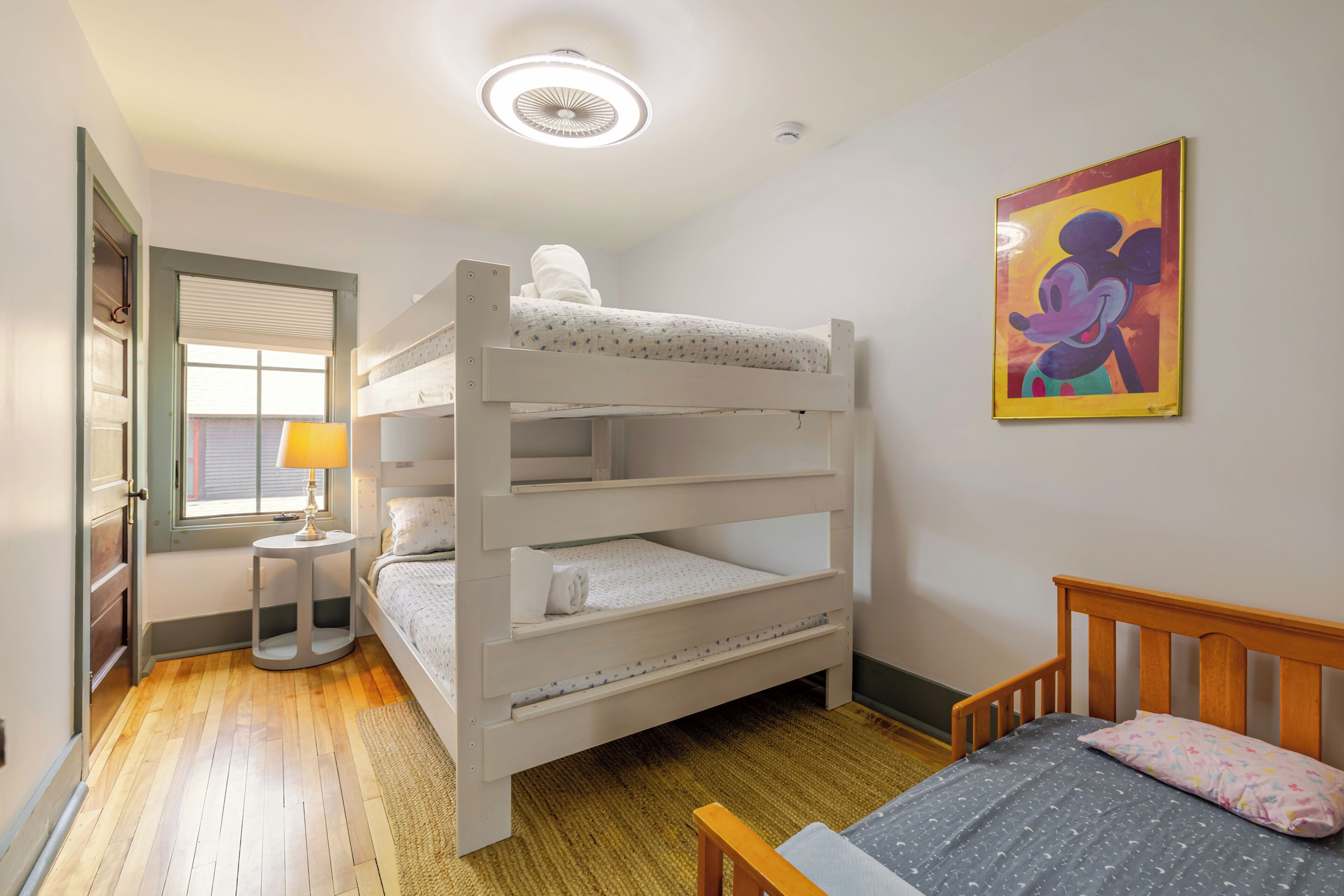 Une chambre avec un lit superposé, une fenêtre, une lampe et un tableau représentant Mickey Mouse.
