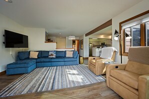 Una sala de estar con un sofá modular, un sillón color arena, un tapete estampado, un televisor de pantalla plana y una cocina al fondo.