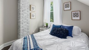 Ein Schlafzimmer mit einer Wand in Ziegeloptik, einem Bett mit weißer Bettwäsche und blauen Kissen, einem Fenster mit weißen Vorhängen und gerahmten Kunstwerken an der Wand.