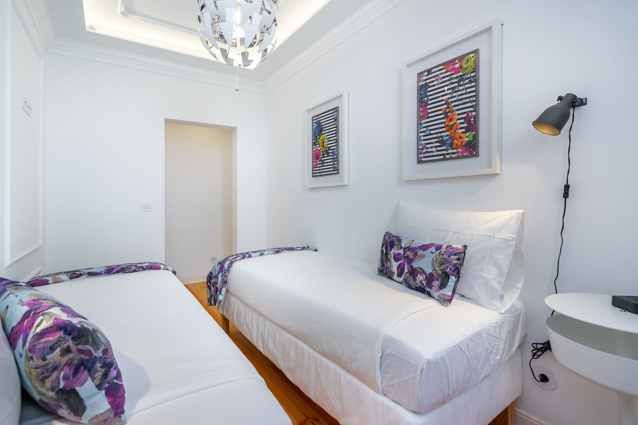Une chambre avec deux lits, un plafond blanc, un lustre, une lampe fixée au mur et une œuvre d’art encadrée.