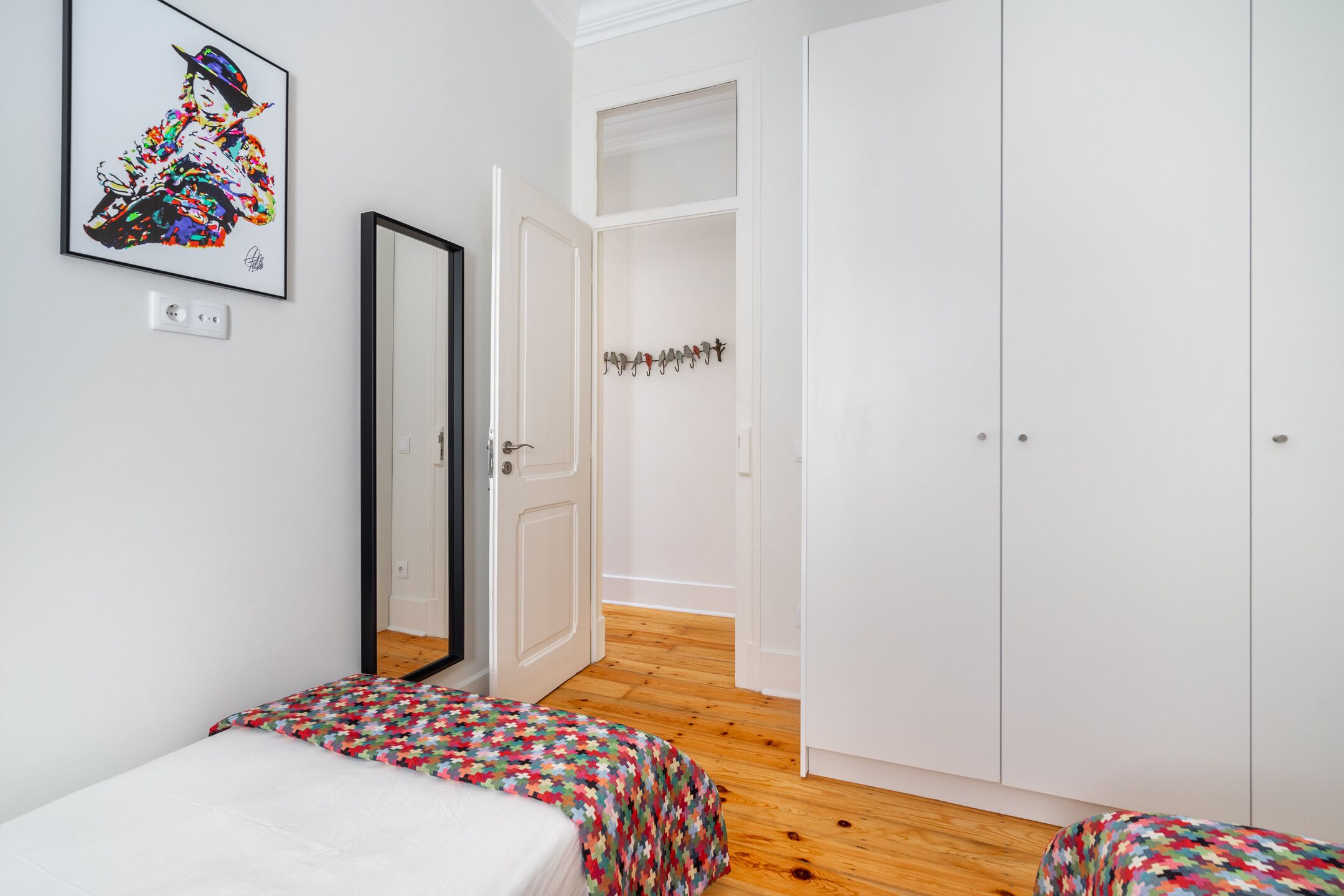 A bedroom with two beds, a wardrobe, a mirror, and a colorful artwork on the wall.