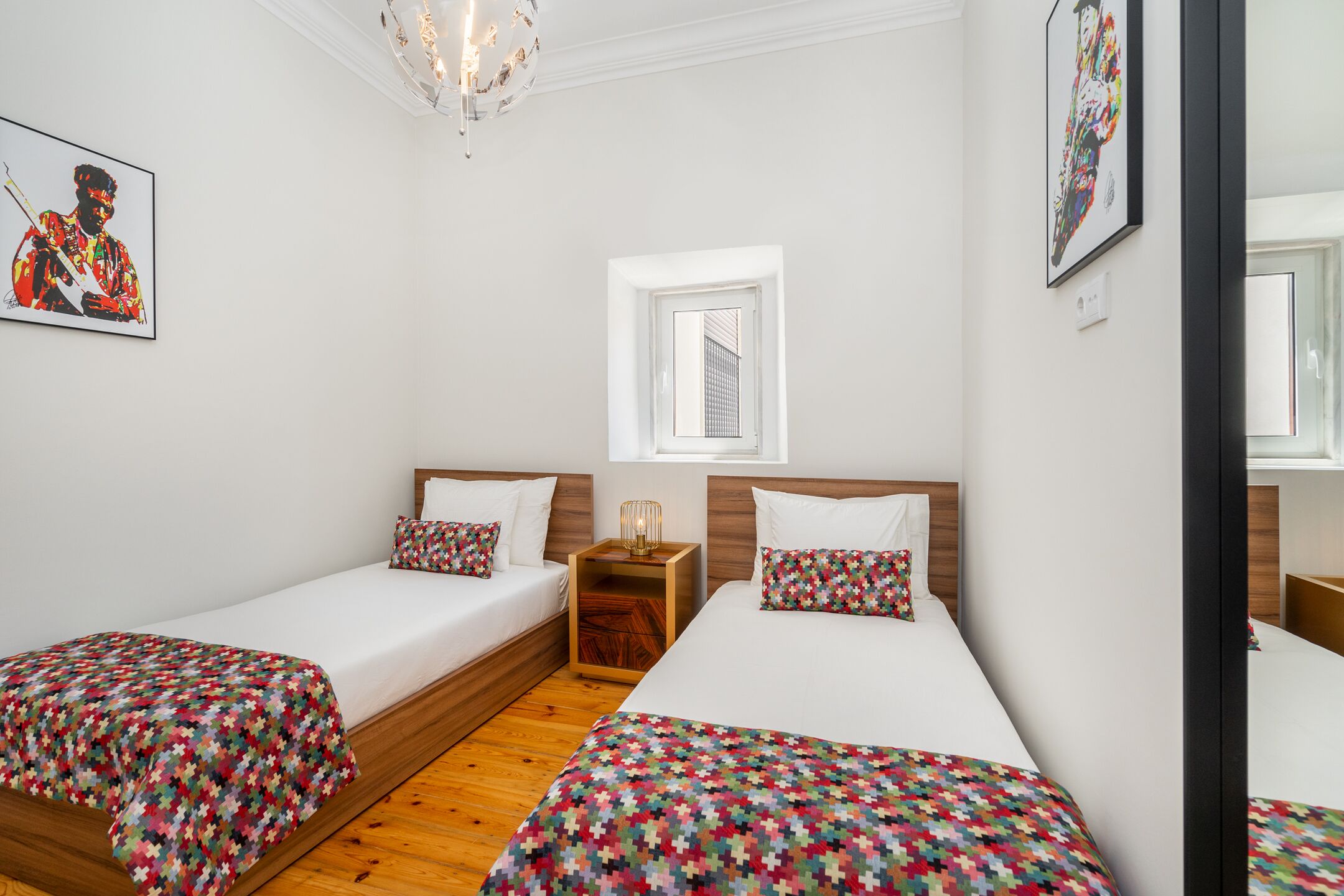 A hotel room with two single beds, a wooden nightstand, a small table, and a window with a view of a building.