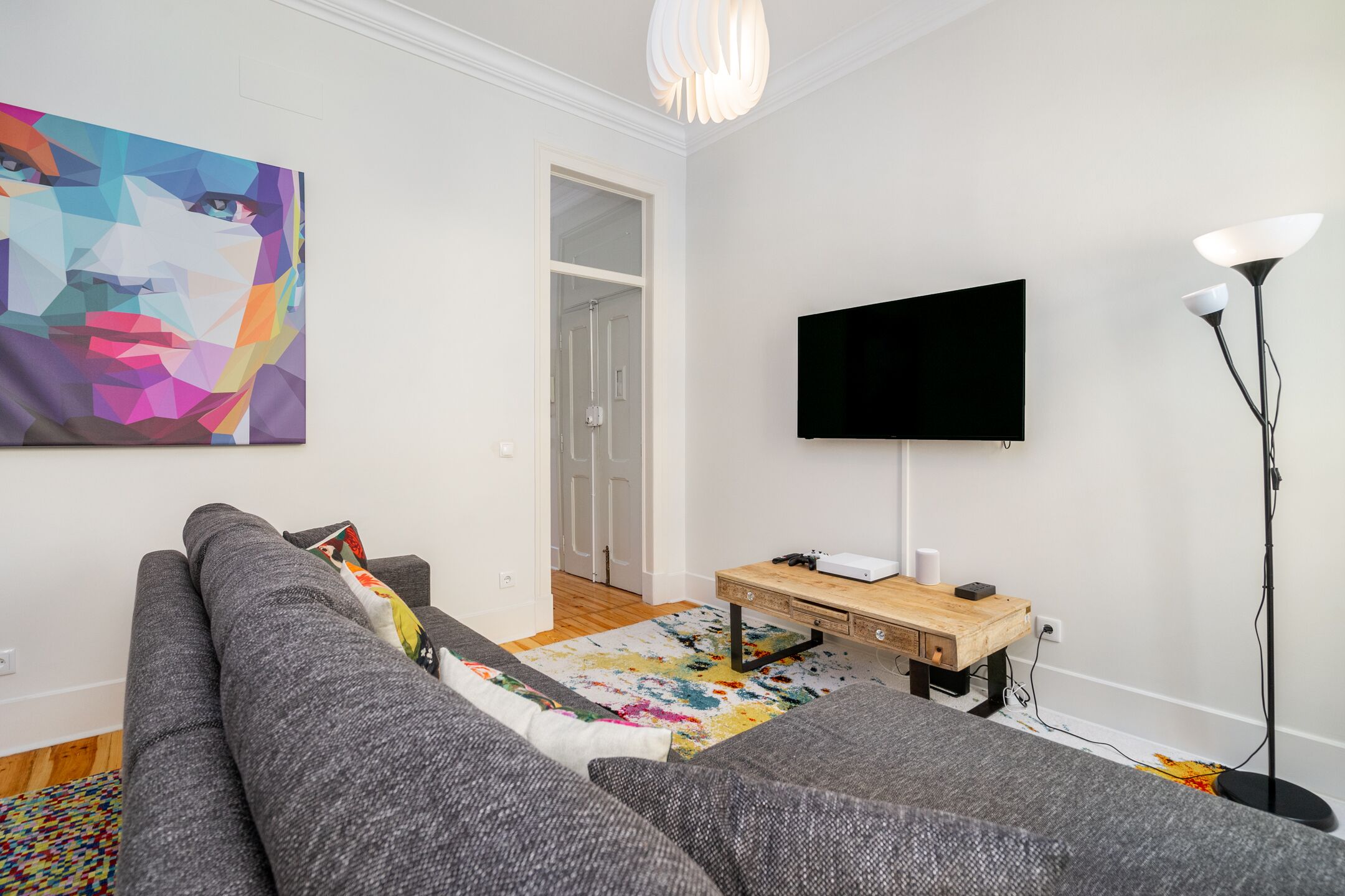 Un salon moderne avec un canapé gris, un tableau abstrait aux couleurs vives, un téléviseur à écran plat, une table d’appoint en bois et un lampadaire.