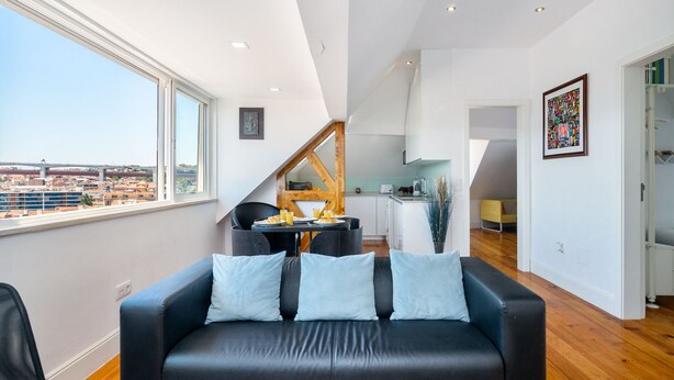 A modern living room with a black leather sofa, a dining area with a wooden table, and a kitchen in the background. - Premium River View Apartment (Lisbon)