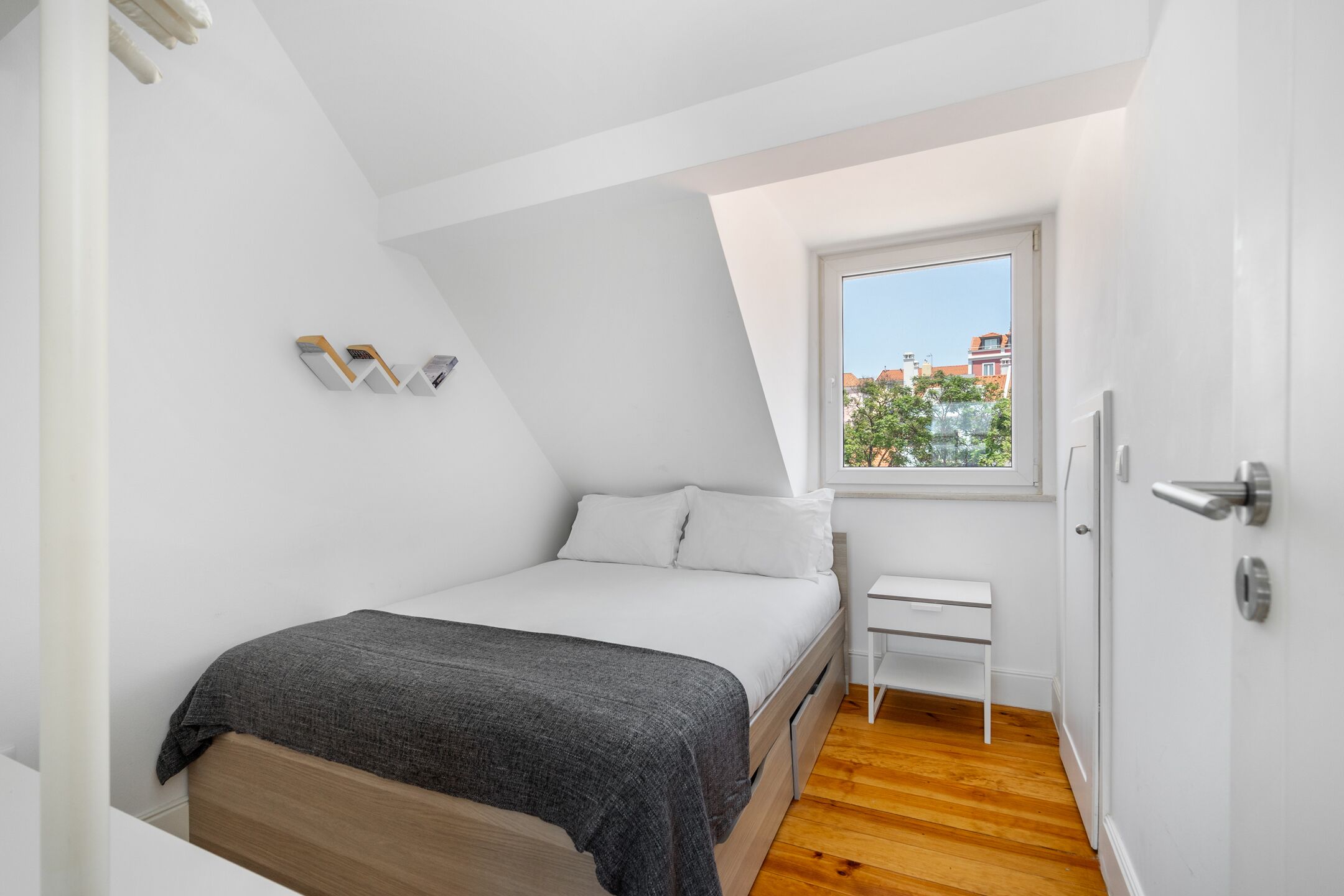 Una camera da letto con un letto, una scrivania piccola e bianca, una finestra con vista sugli edifici e una porta.