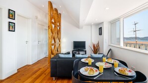 A modern living room with a black sofa, dining table with plates of food and drinks, a TV, and a large window with a view. - Premium River View Apartment (Lisbon)