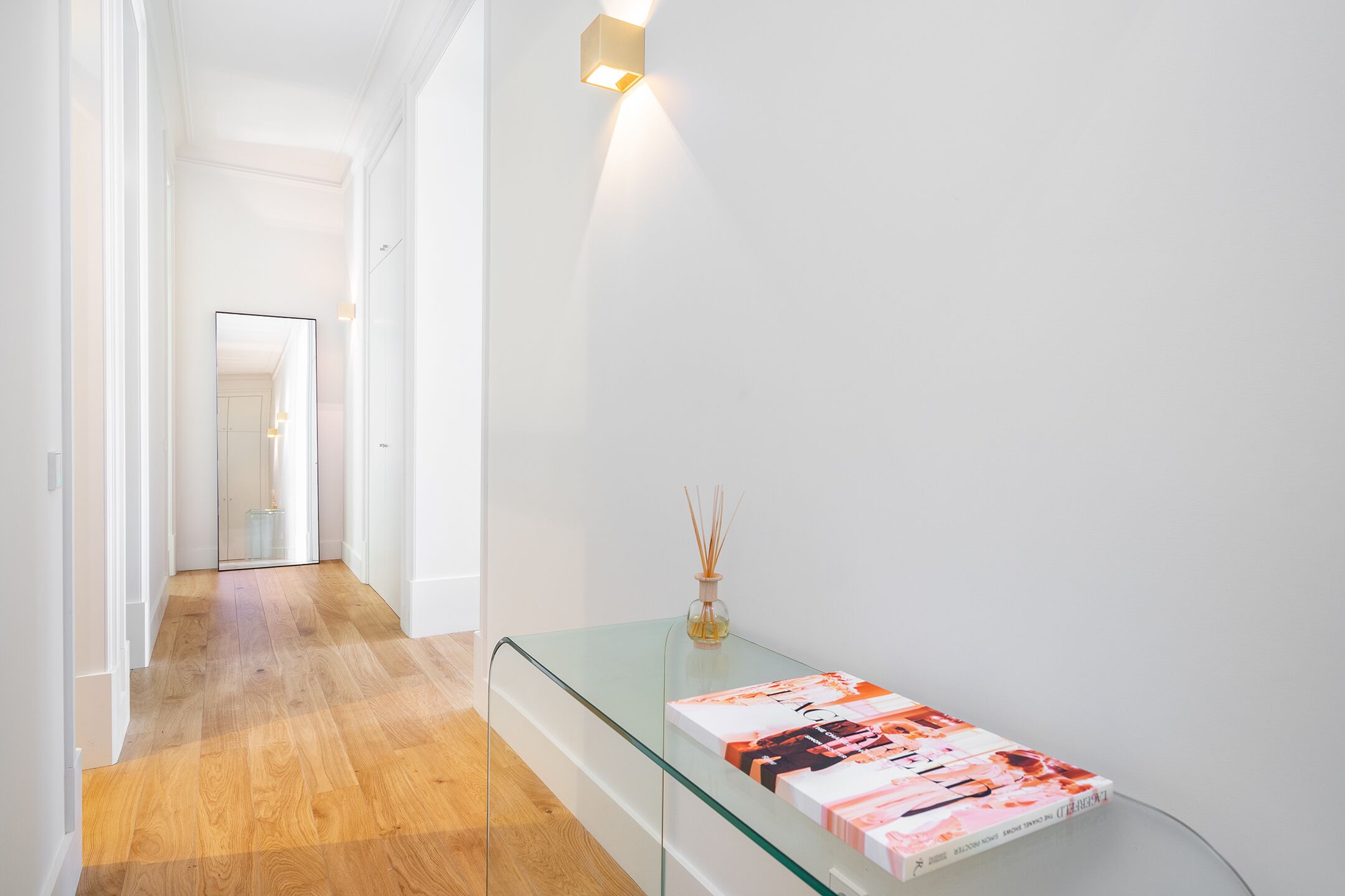 A hallway with a glass console table displaying a magazine, a wall-mounted light, and a wooden floor.
