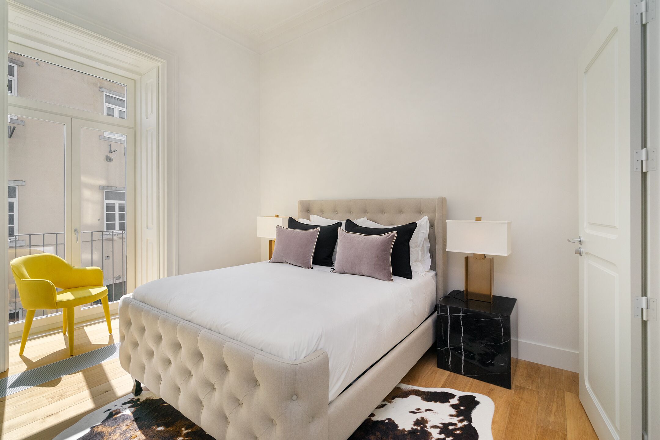 A bedroom with a tufted headboard bed, a nightstand with a lamp, a yellow chair, and a view of the outside balcony.