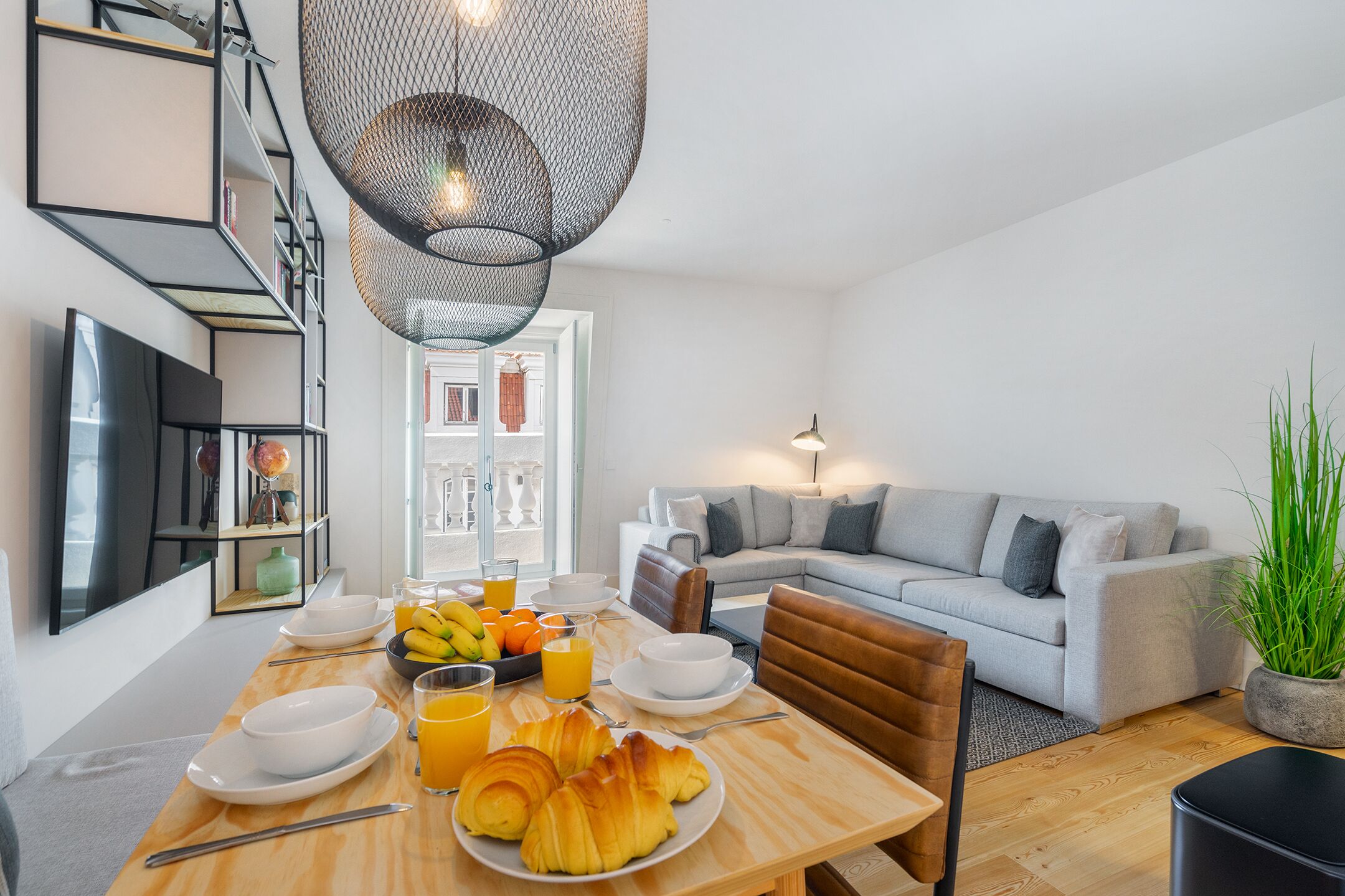 A modern living room with a grey sofa, a dining table set with breakfast, and a large window.