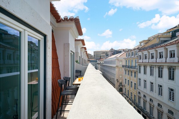 A narrow balcony with a long table and chairs, overlooking a row of buildings with classic architecture.