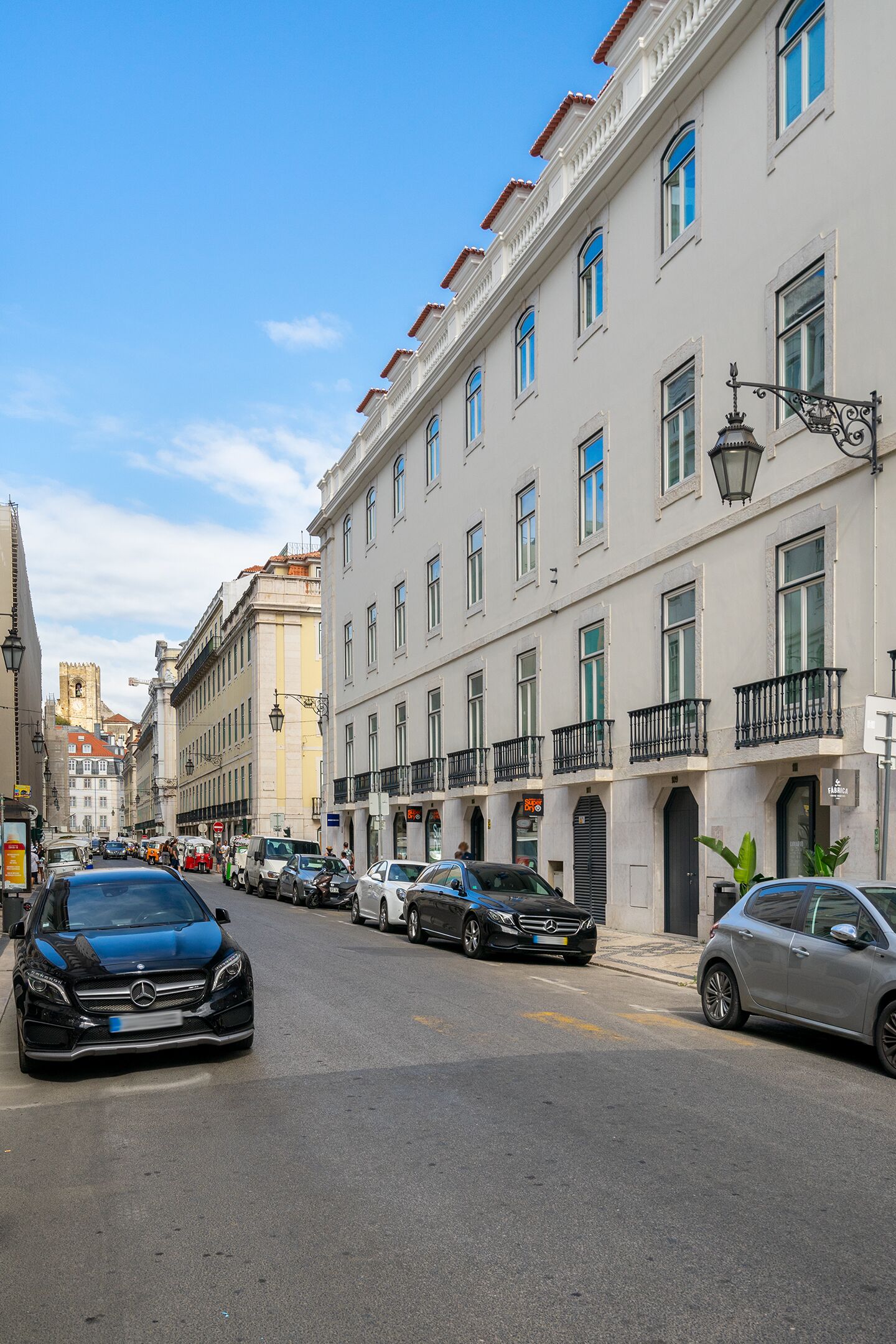 Rua com edifícios altos, carros estacionados e céu azul.