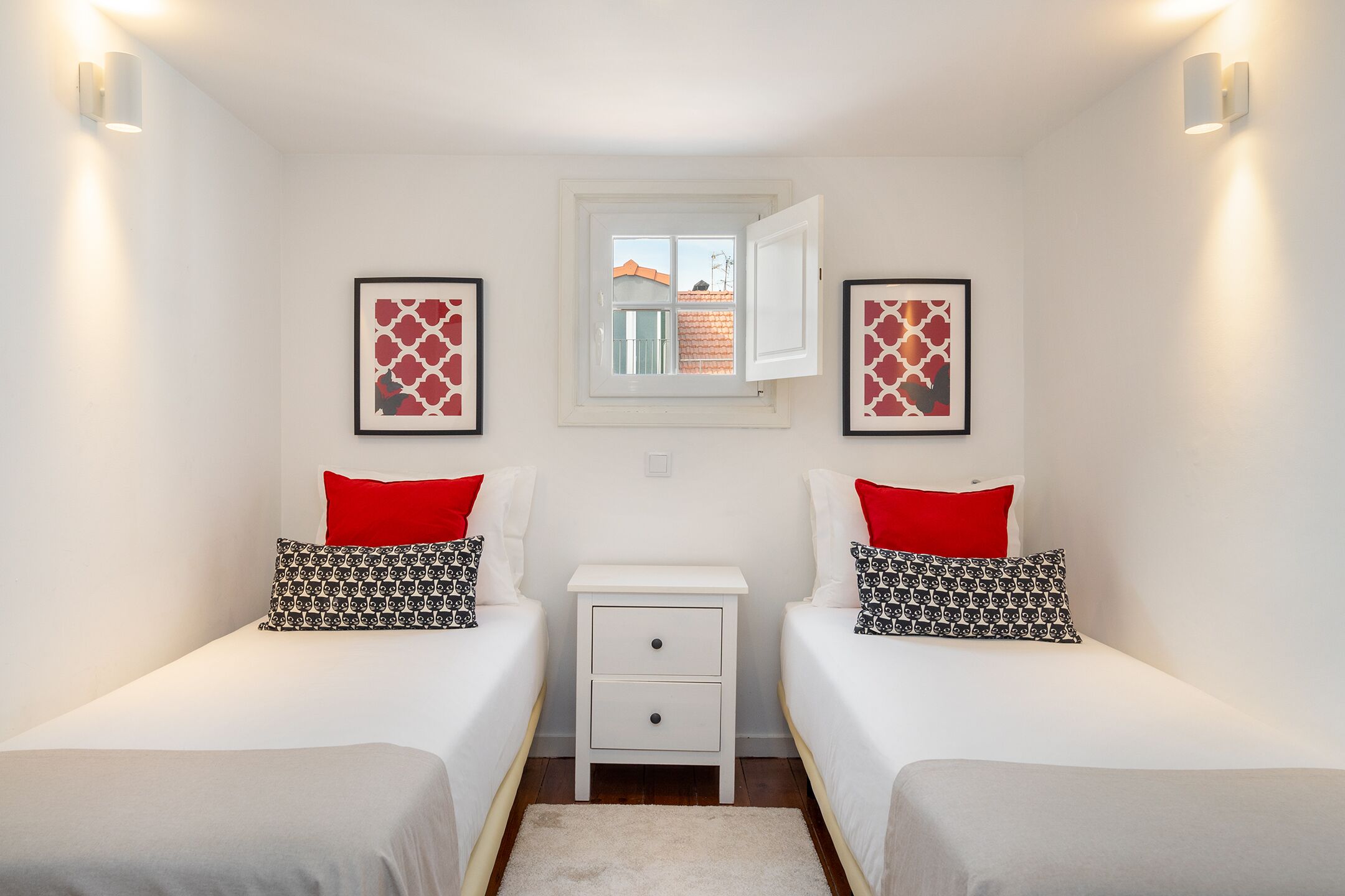 A twin bedroom with two beds, a small white nightstand, a window with an open shutter, and framed artwork on the walls.