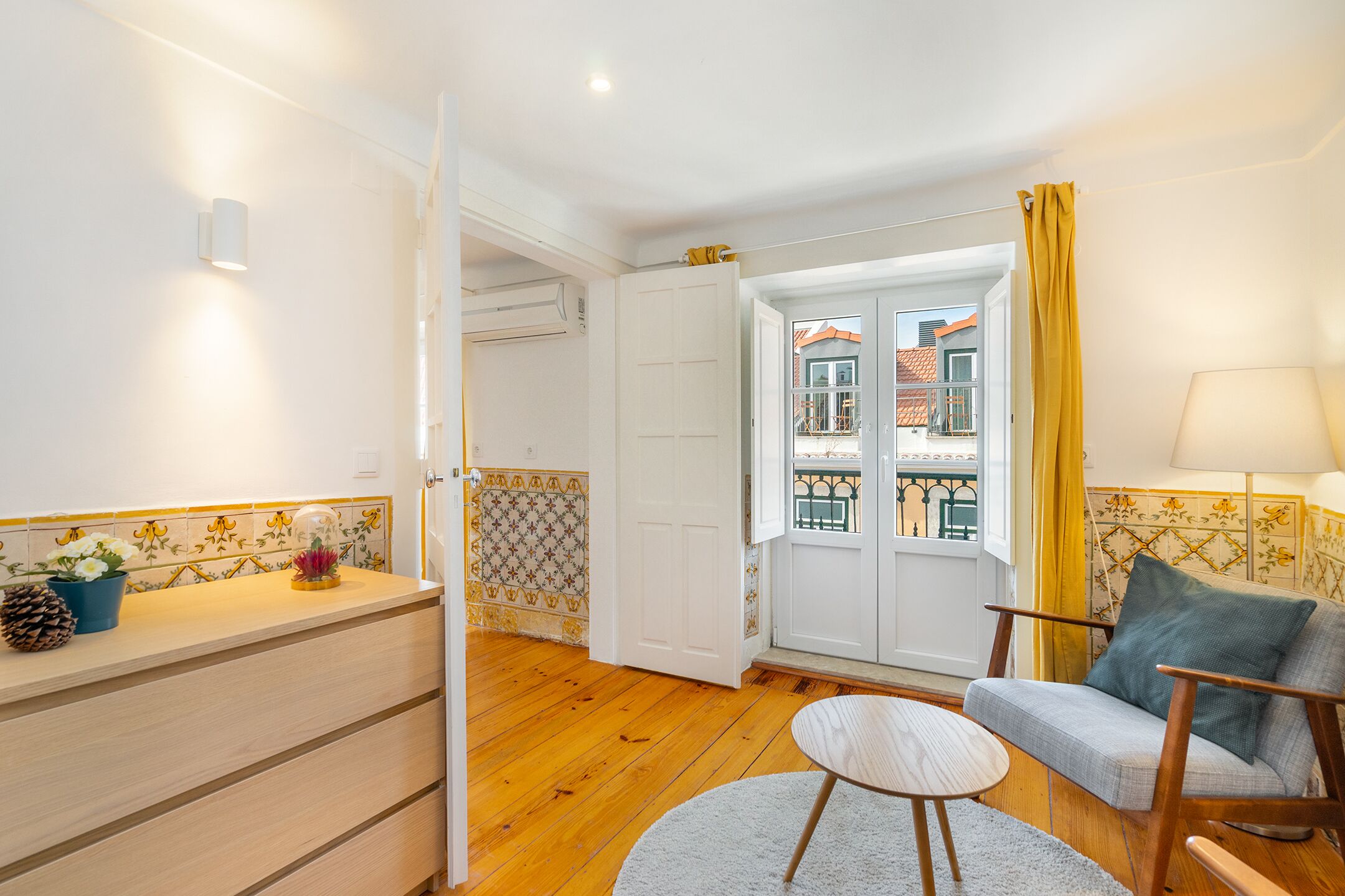 A room with a wooden floor, a white door, a window with yellow curtains, a sofa, and a small table.