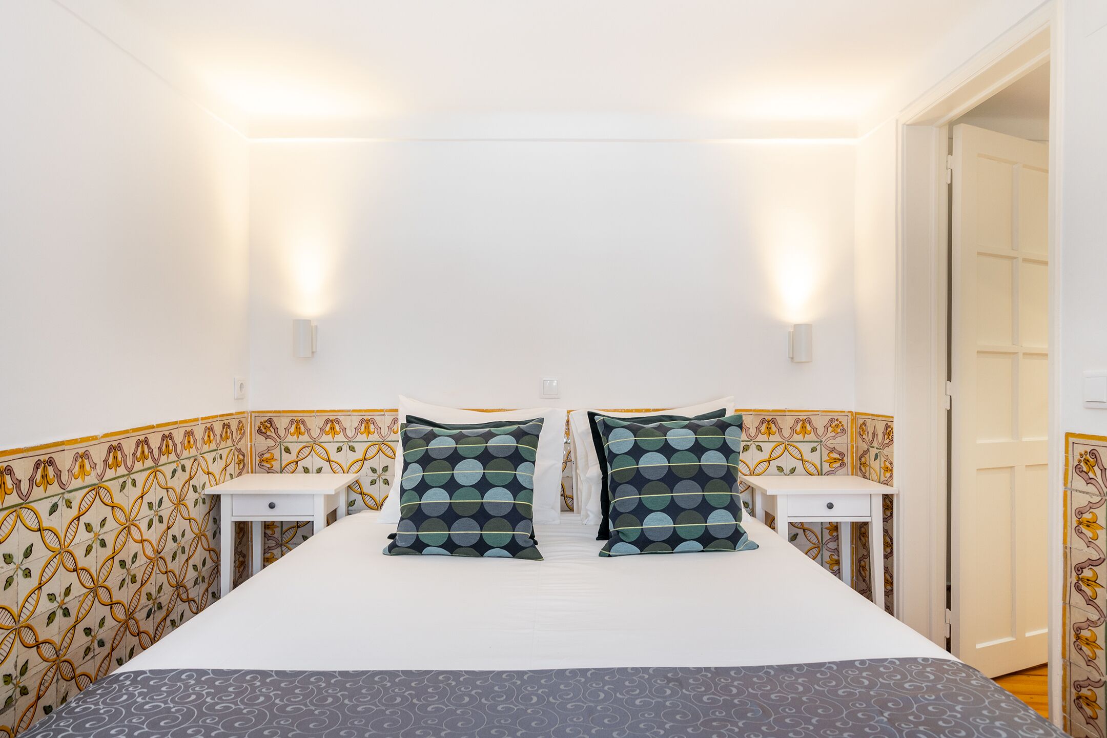 Une chambre d’hôtel avec un lit, deux tables de chevet, des carreaux muraux à motifs et une porte.