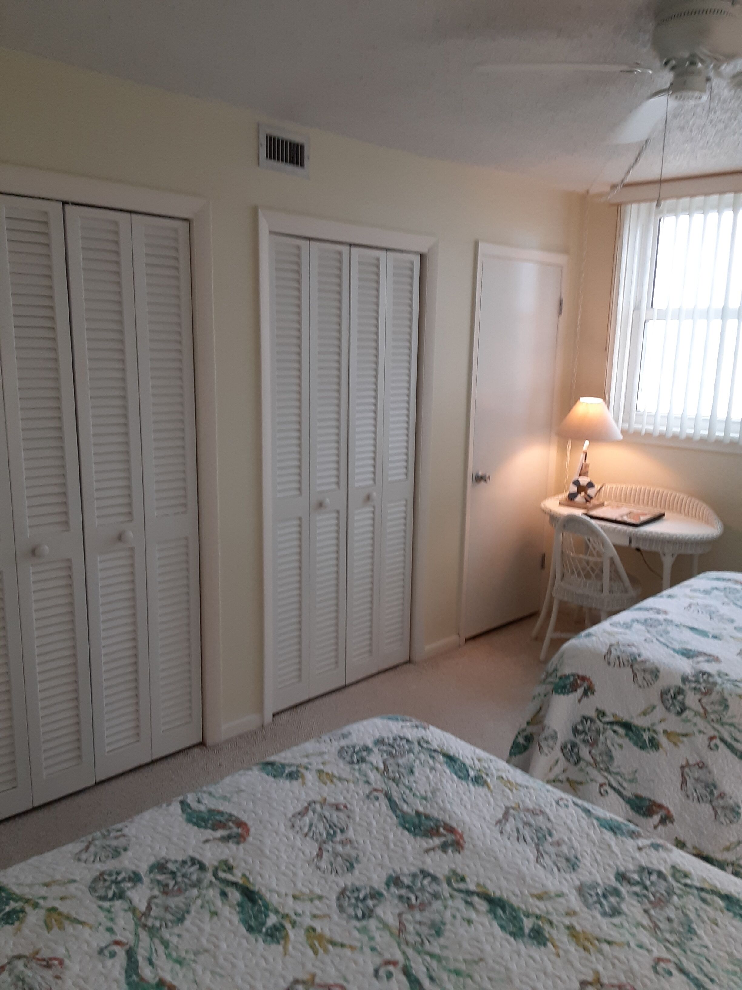 Une chambre avec deux lits, un ventilateur de plafond blanc, une fenêtre avec des stores et une petite table avec une lampe.