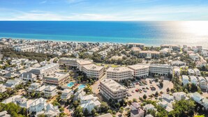 An aerial view of a coastal town with a mix of residential and commercial buildings, a clear blue sea, and a bright sun.
