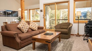 Ein geräumiges Wohnzimmer mit einem braunen Sofa, einem Holztisch und Blick durch große Schiebeglastüren auf eine Terrassenanlage.