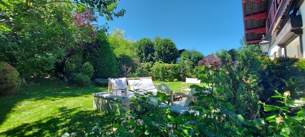 Relax in a serene garden surrounded by lush greenery and flowering plants, with a stone bench and neatly trimmed hedge.