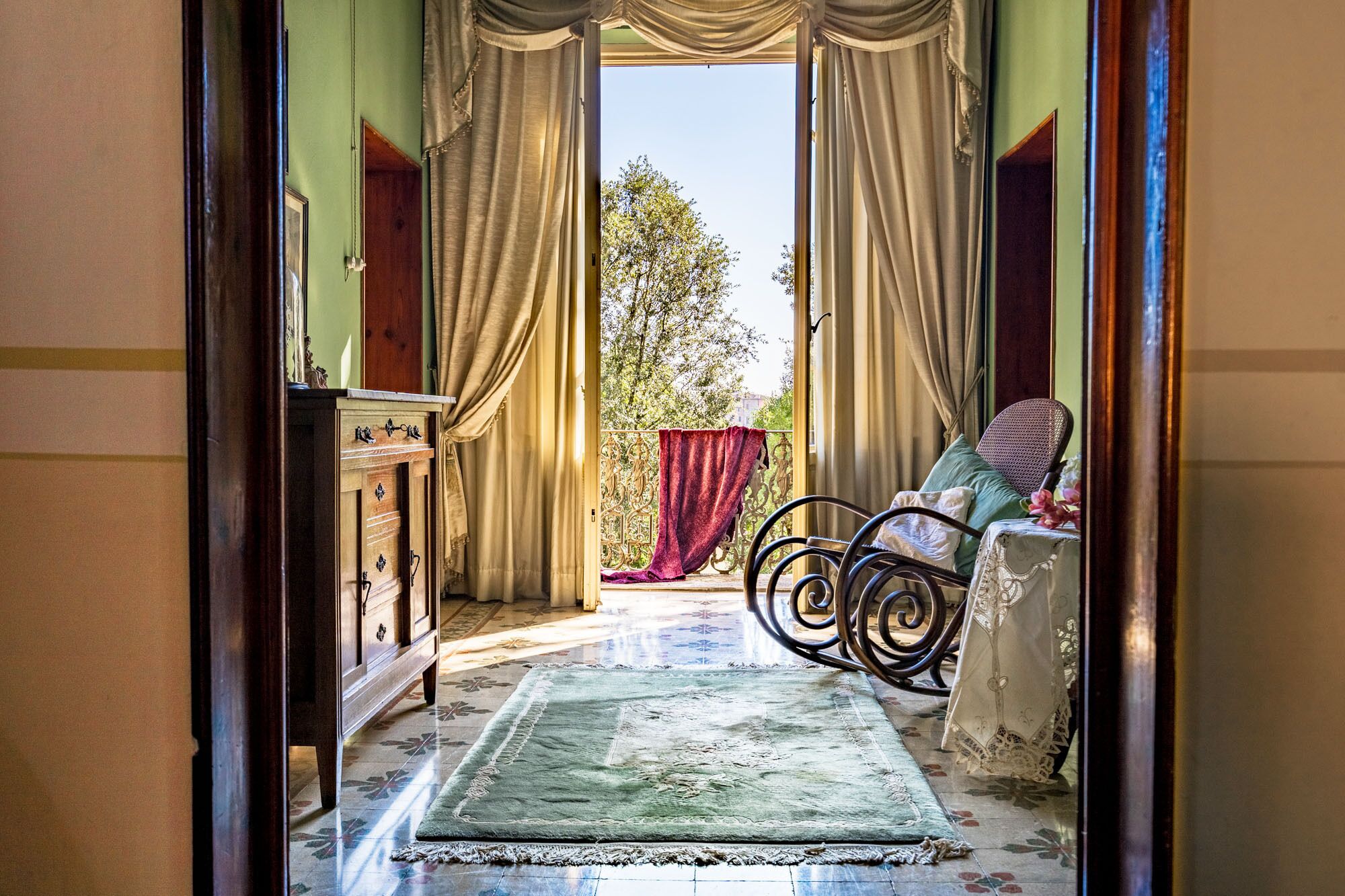A room with a view of trees through open curtains, a patterned rug, a chair with a cushion, and a wooden dresser.