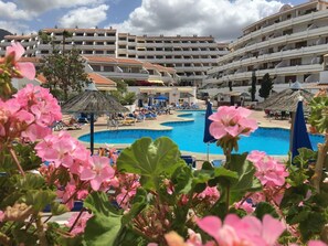 A hotel with a swimming pool surrounded by lounge chairs and umbrellas, a large building with multiple balconies, and tropical flowers in the foreground. - Studio at Garden City, Costa Adeje - Heated  Pool and pool bar  Near sandy beach (Playa las Americas)