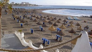 Ein Strand mit Reihen aus Strohdächern und blauen Liegestühlen, freier Blick auf das Meer und eine entfernte Küstenlinie mit Gebäuden.