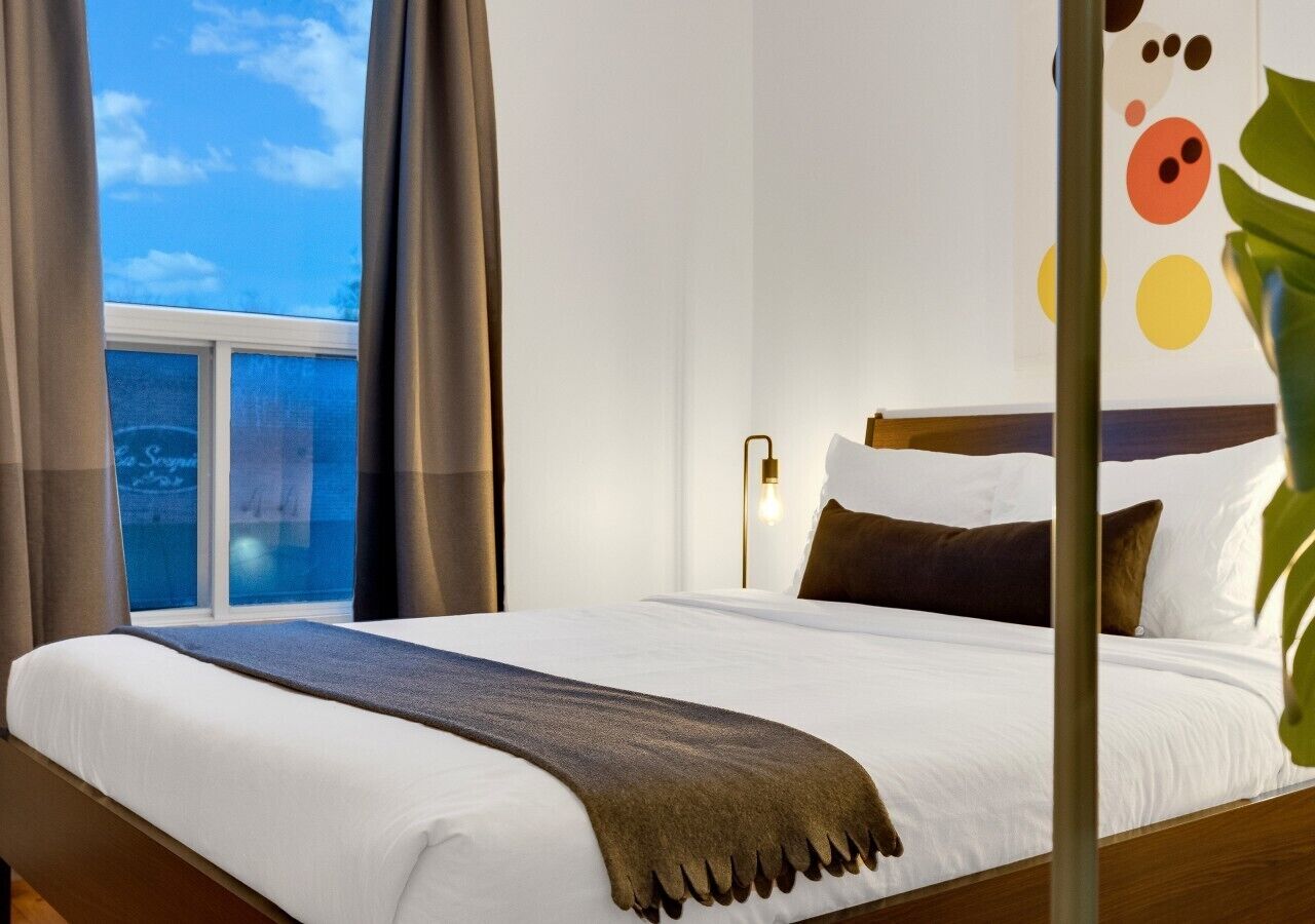 A hotel room with a large bed, a window with curtains, a wall-mounted light, and a partial view of a plant.