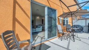 Une terrasse ensoleillée avec une porte-fenêtre coulissante donnant sur un espace de vie, un coin salon extérieur et une table abritée.