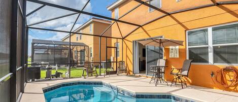 A swimming pool with a glass enclosure, outdoor furniture, and a view of a yellow building.