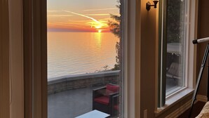Huoneessa on suuri ikkuna, josta avautuu näkymä auringonlaskuun veden yllä, pieni patio, jossa on tuoli, sekä seinässä oleva lamppu.