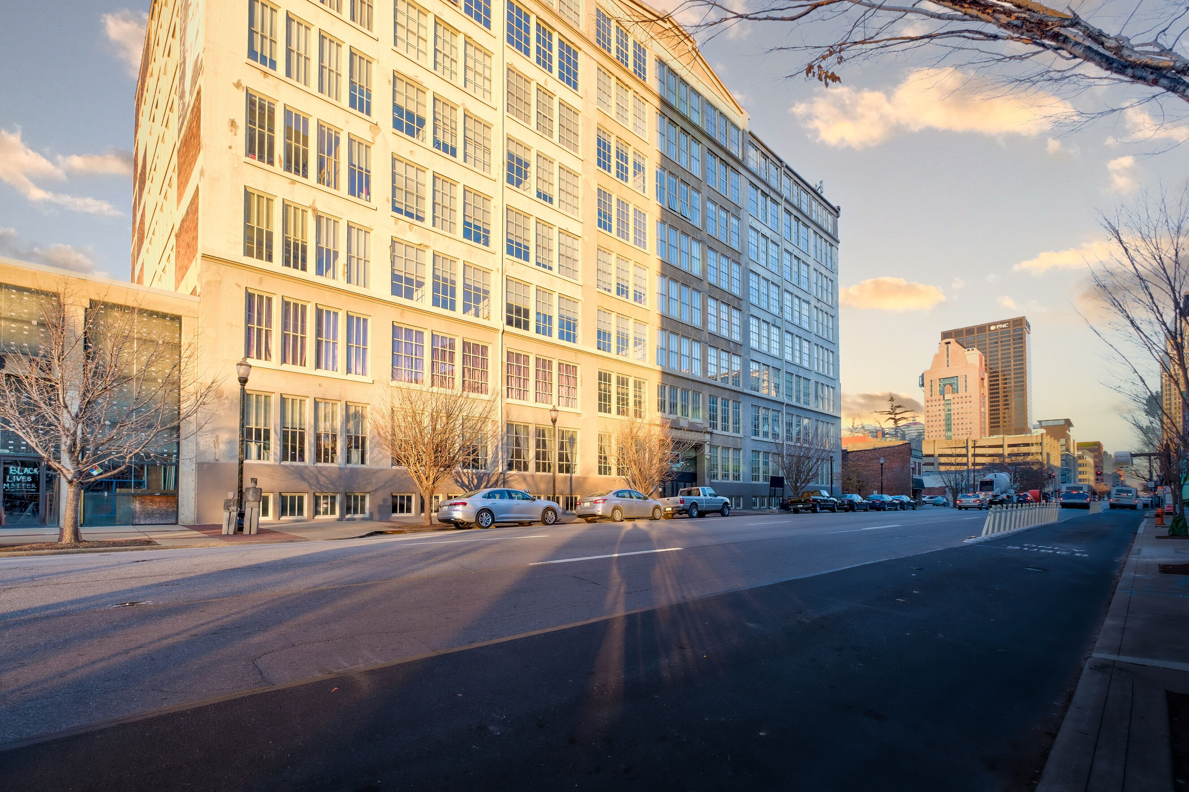A multi-story building with large windows and a flat facade, located on a city street with parked cars and a clear sky.