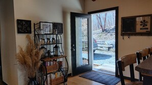 A cozy interior with a wooden floor, a black metal shelf with books and decorative items, a door leading outside, and a dining area with chairs. - Claytor Lake Hideaway (Pulaski)