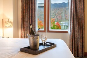 A hotel room with a bed, a bedside table with a bottle of champagne and two glasses, and a window with a view of autumn foliage. - 2 BR Lodge at Spruce Peak (Stowe)