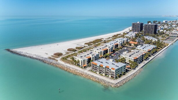 Vista aérea de un complejo turístico frente al mar, con propiedades en primera línea de playa, una playa de arena y un mar azul y claro.