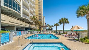 A pool area with multiple pools, lounge chairs, palm trees, and a building with balconies. - Ocean Villa 1302 (Panama City Beach)