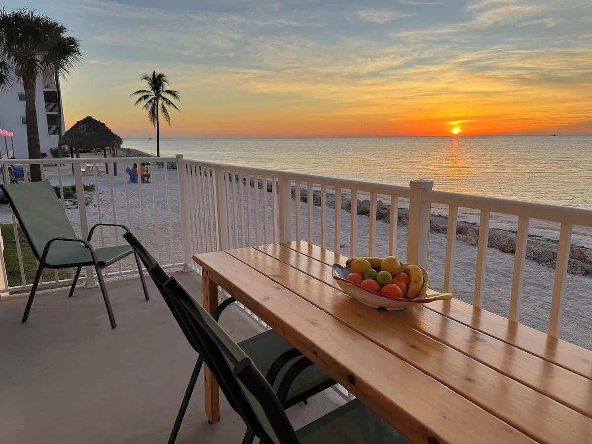 A wooden table with a bowl of fruit on a balcony overlooking the sea at sunset.