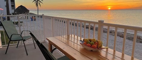 Et træbord med en skål frugt på en balkon med udsigt over havet i solnedgangen.