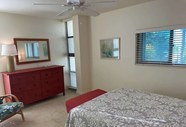 A bedroom with a bed, a red dresser, a ceiling fan, and two windows with blinds.