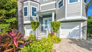 Uma casa moderna de dois andares, com varanda frontal coberta, garagem e um jardim bem cuidado com plantas tropicais.