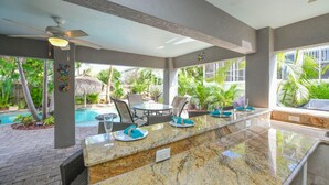 Um bar à beira da piscina com bancada de mármore, mesas ao ar livre e vista para a piscina e plantas tropicais.