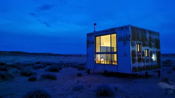 Un petit bâtiment de forme cubique, doté de grandes fenêtres, situé dans un paysage désolé, au crépuscule.